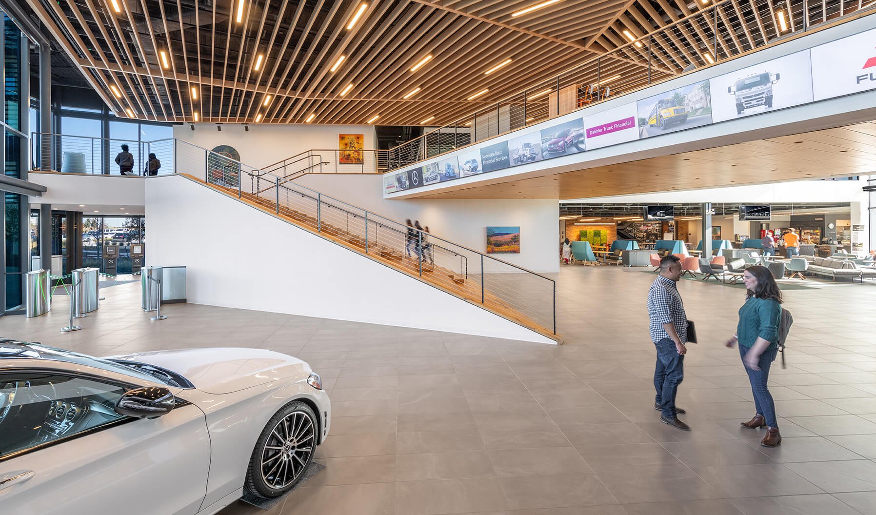 Upon entering the building, you are introduced to an impressive open space showcasing a Mercedes-Benz vehicle. Just beyond the staircase is a large gathering area with a cafeteria perfect for collaboration or individual relaxation. 