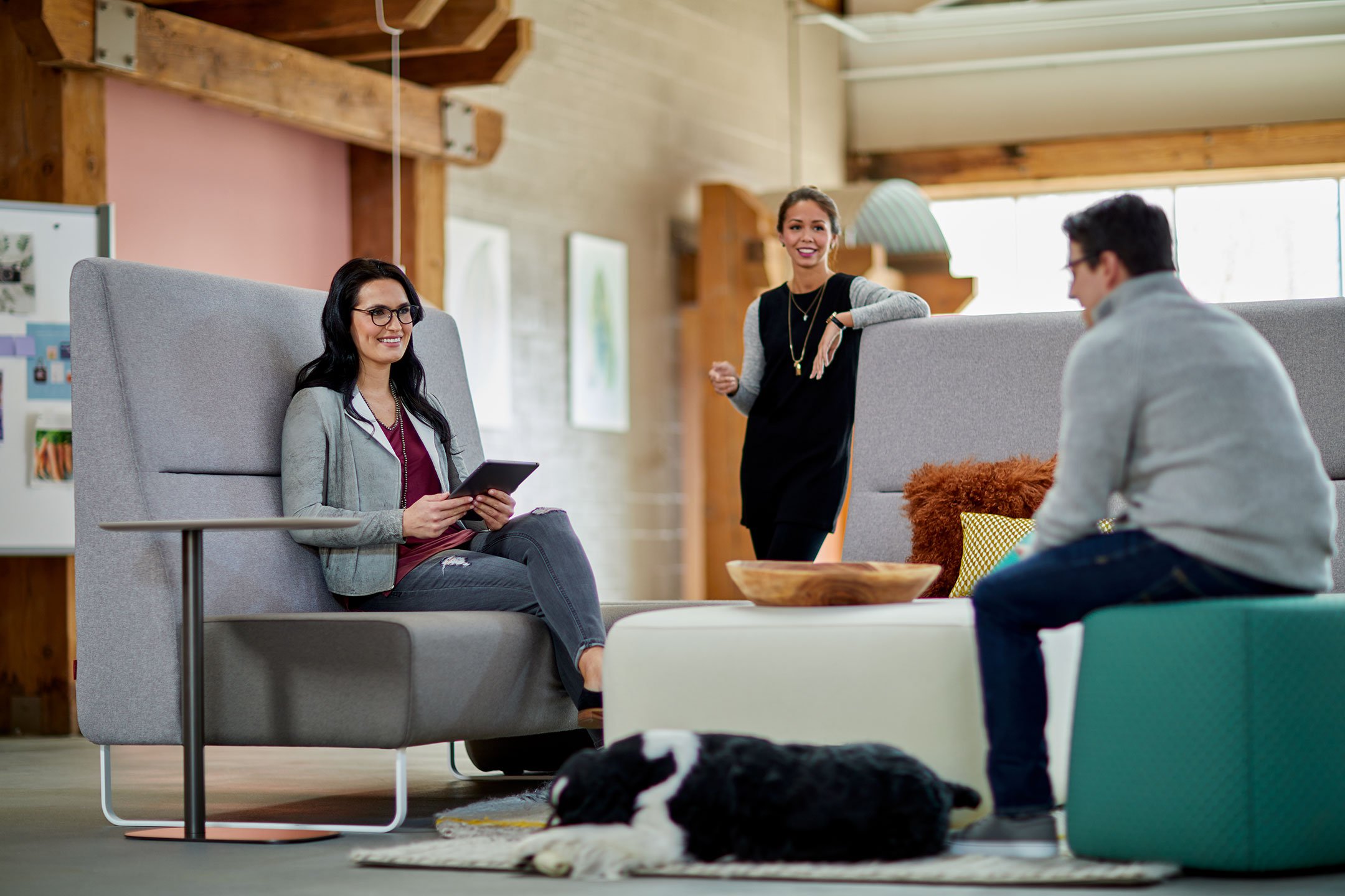 Haworth Riverbend and Pebble Booth in grey color with white and green ottoman and employees talking in the space with a dog laying next to couch 